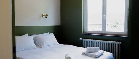 A hotel room with a bed, white linens, a window with a view, a radiator, and a wall-mounted light fixture.