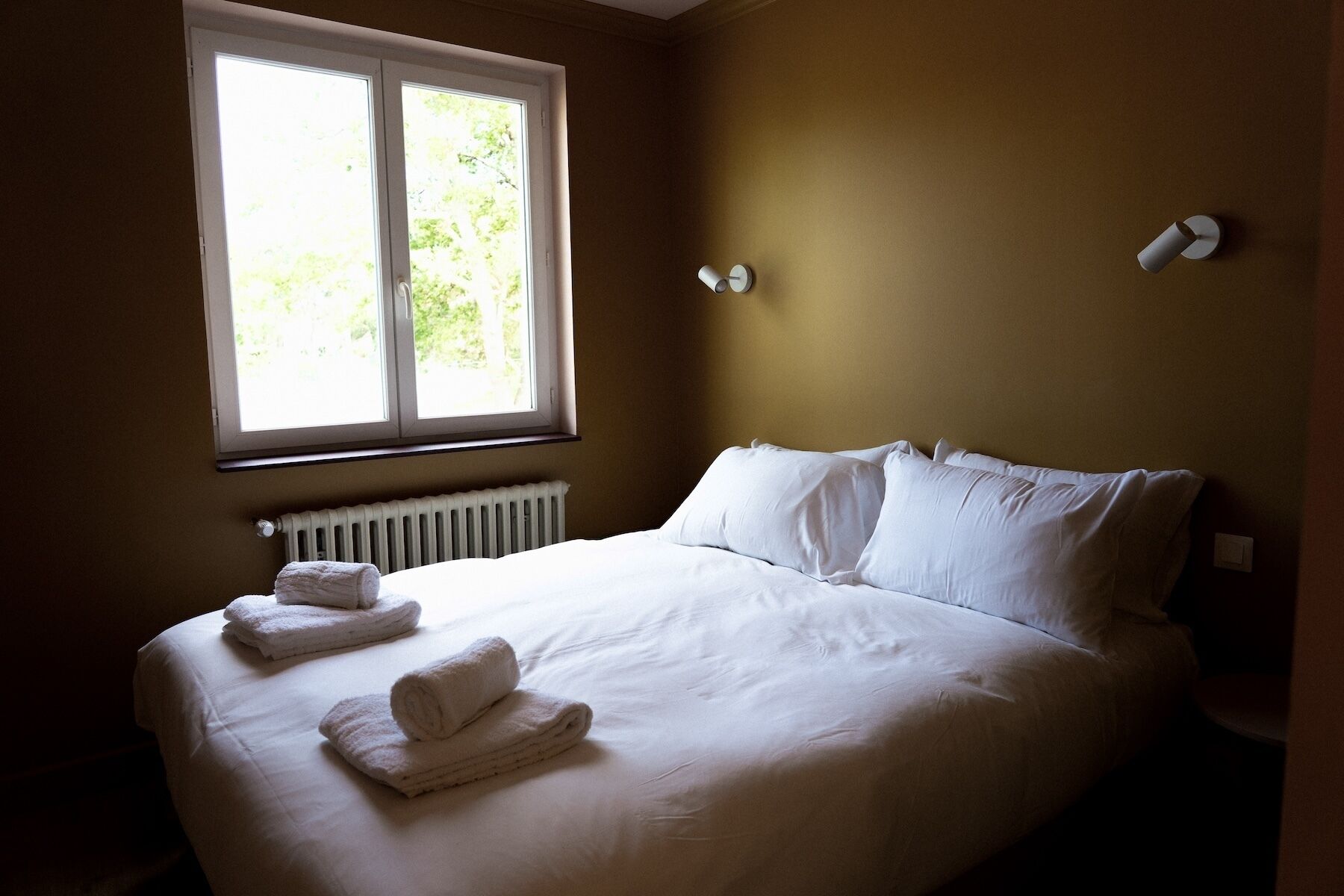 A neatly made bed with white linens and two rolled towels, a window with a view of greenery, and a wall-mounted light fixture.
