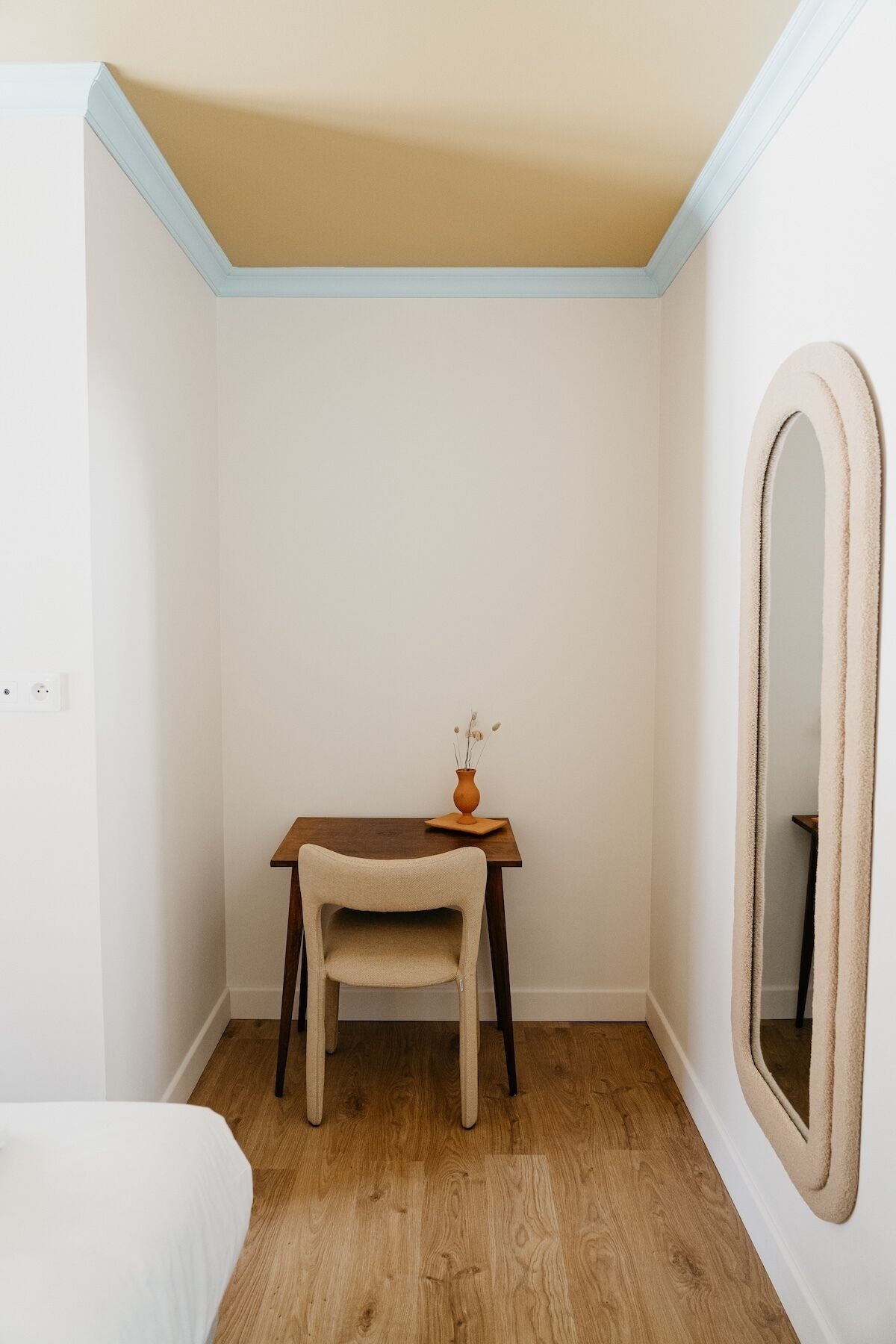 Une petite chambre à coucher, bien rangée, avec un bureau en bois, une chaise, une petite table sur laquelle est posé un vase, et un grand miroir.