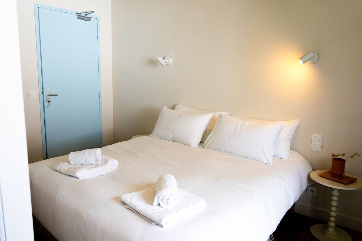 A neatly made bed with white linens and two rolled towels on the bed.