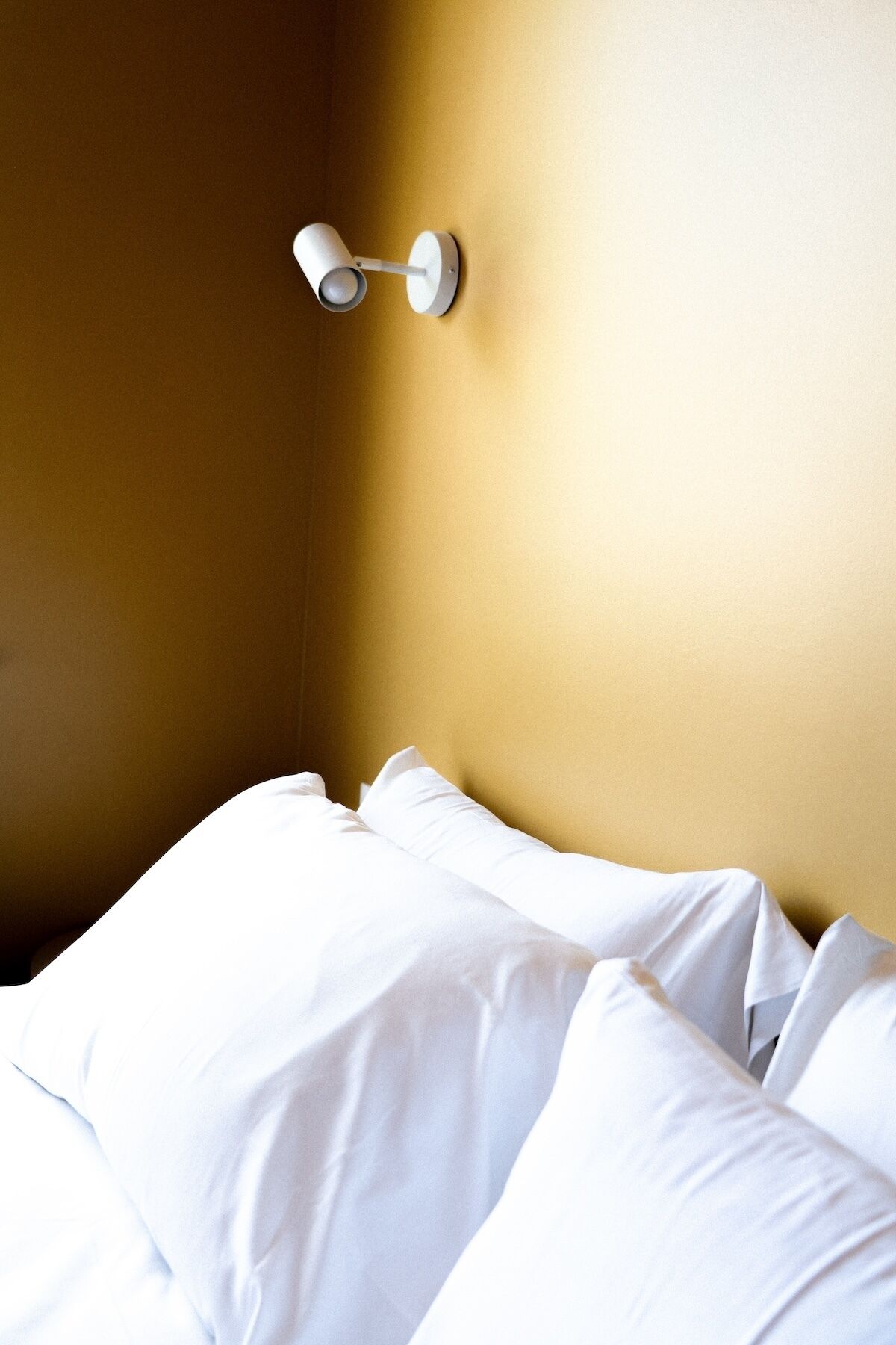 A hotel room with a bed against a yellow wall, a white pillow, and a wall-mounted light fixture.