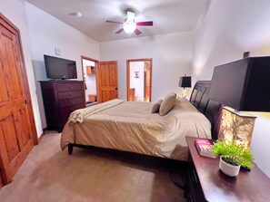 Un dormitorio con una cama, un televisor sobre una cómoda, un ventilador de techo, puertas de madera y una pequeña planta en maceta.