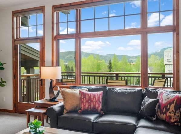 A room with a black leather sofa, a lamp, and a view of green hills through large windows.