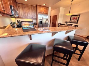 A kitchen with granite countertops, a stainless steel refrigerator, wooden cabinets, and a laptop on the counter. - Timberline Cove Lakeside Condo • Mtn Views • Hot Tubs • Bike Path (Frisco)