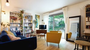A modern living room with a sofa, yellow armchair, and a bookshelf filled with books. A television is mounted on the wall, and there is a large window providing a view of the outdoors. - Quiet family home with terrace - near Ondres beach (Ondres)