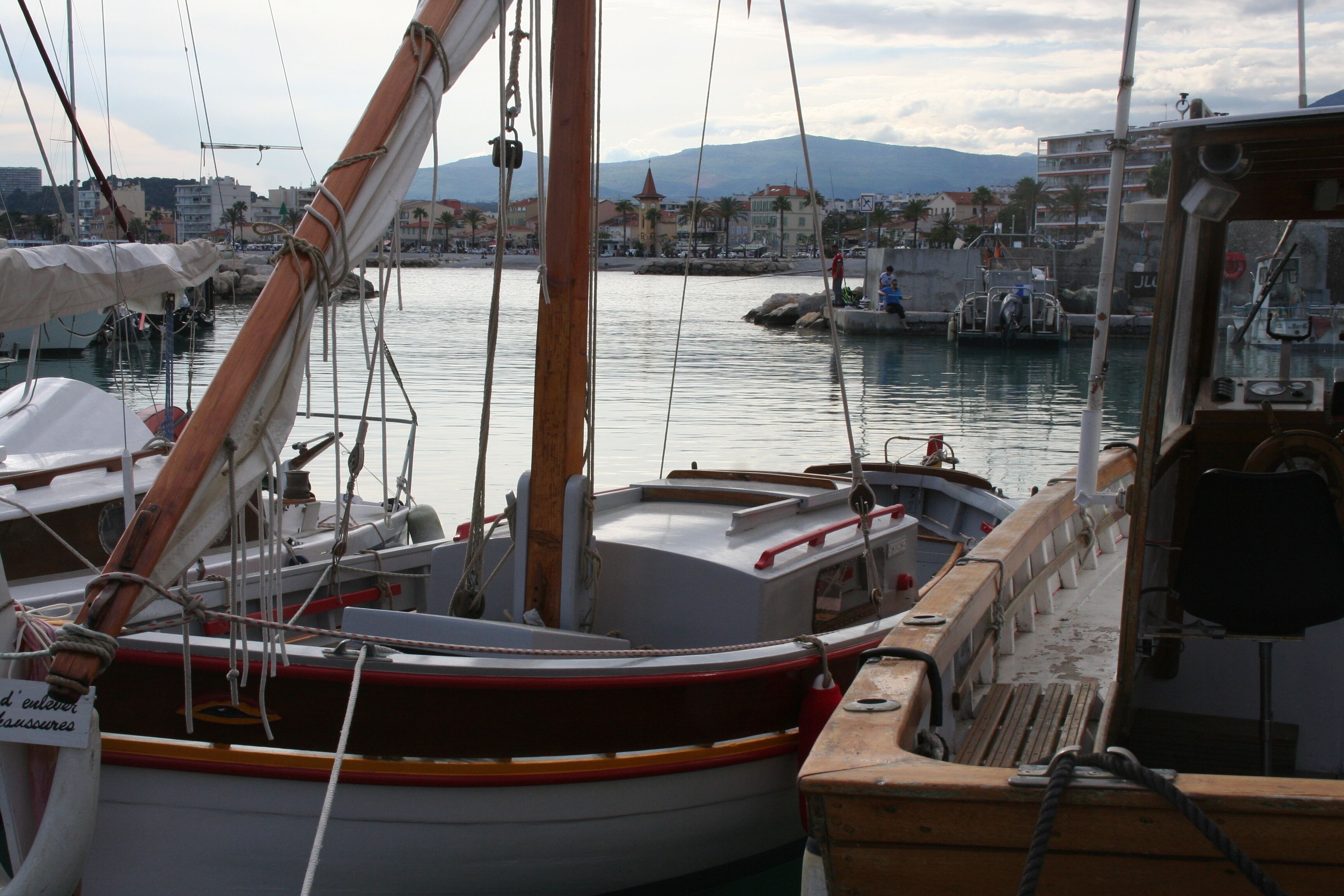 Un puerto deportivo con barcos amarrados, un paisaje urbano al fondo y una línea de montañas en el horizonte.