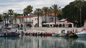 Un port de plaisance avec des bateaux amarrés, un bâtiment en pierre doté d’arcades et des palmiers.