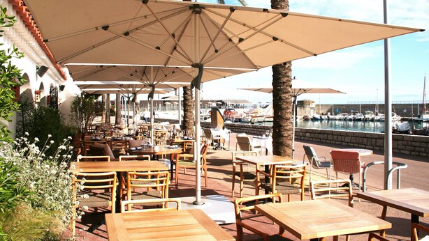 Un espace extérieur aménagé avec des tables et des chaises en bois, de grands parasols et une vue sur une marina où sont amarrés des bateaux.