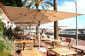 An outdoor seating area with wooden tables and chairs, large umbrellas, and a view of a marina with boats.