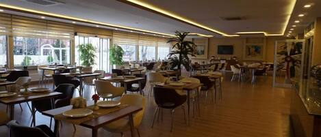 A spacious restaurant with multiple tables and chairs, large windows, and indoor plants.