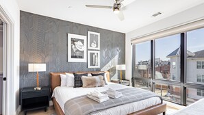 Ein modernes Schlafzimmer mit einem großen Bett, einem Deckenventilator und Blick auf die Stadt durch große Fenster.