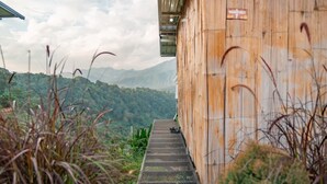 A rustic wooden building with a metal staircase leading to a platform, surrounded by lush greenery and mountains in the background. - Long hug khao homestay (Pong Yaeng)