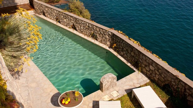 A private pool with lounge chairs and a table, overlooking a coastal sea.