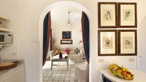 A hotel room with a kitchenette, a dining area with a fruit bowl, and a living area with a sofa and a TV.