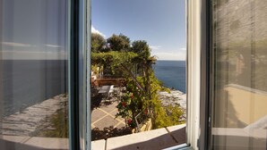 A view from a window overlooking a terrace with outdoor seating, a garden with red flowers, and a clear view of the sea.