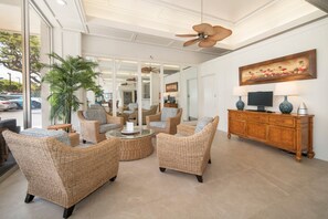 A lobby with wicker furniture, a coffee table, a television on a wooden cabinet, and a large window with a view of parked cars.