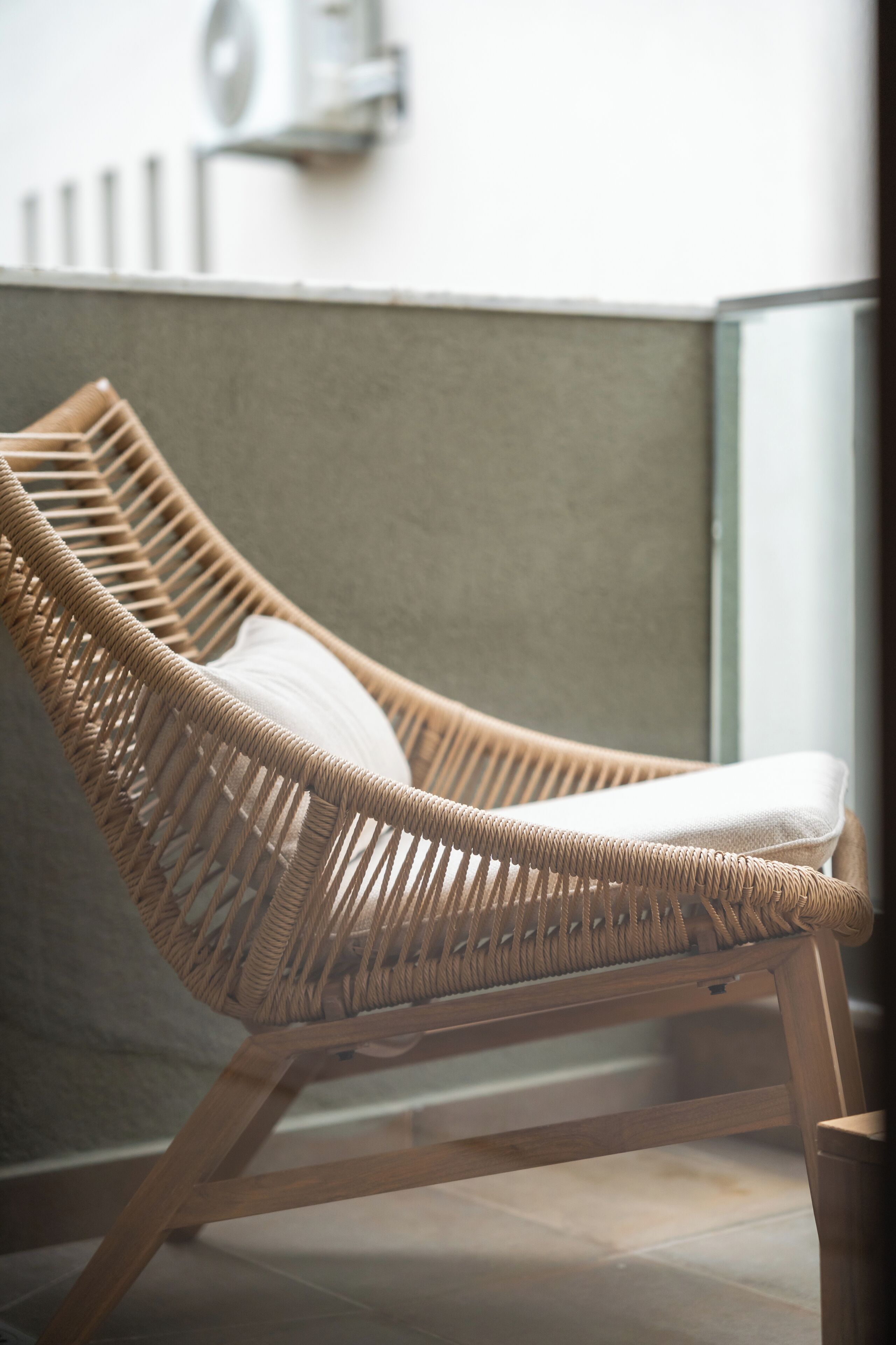 A wicker lounge chair with a white cushion, positioned against a wall with a window.