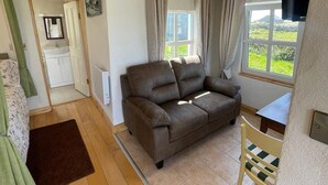 A cozy living room with a brown sofa, a small table, and a window with curtains. - Calla Chalet by the Sea Roundstone (Roundstone)