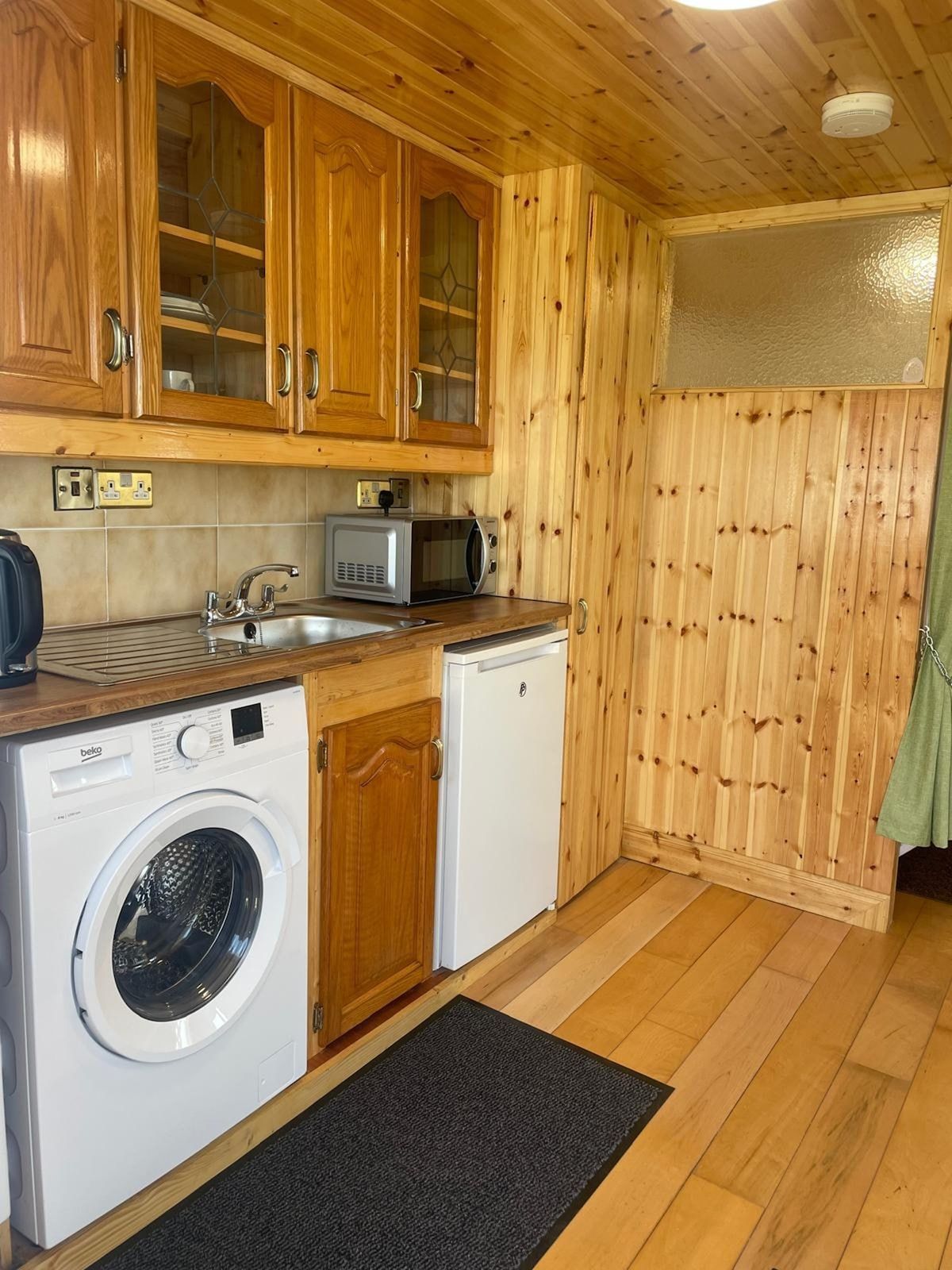 A compact kitchen with wooden cabinets, a washing machine, a microwave, and a small sink.