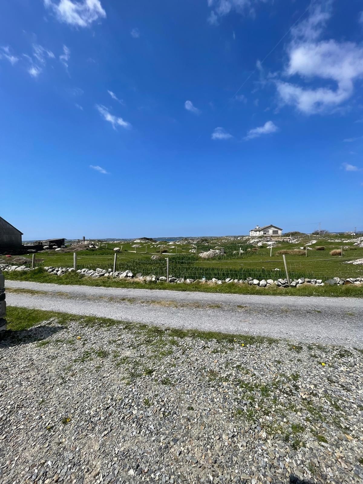 A rural landscape with a gravel road, a stone wall, and a house with a fenced yard.