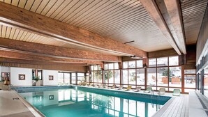Ein Innenpool mit Holzbalken, großen Glasfenstern und Liegestühlen am Beckenrand.
