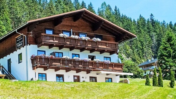 A multi-story wooden building with balconies, set against a backdrop of trees and a clear sky.