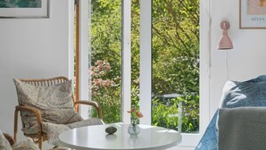 A room with a wicker chair, a round table, a vase with flowers, and a large window with a view of greenery.