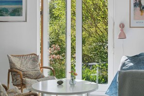 Ein Zimmer mit Korbsessel, rundem Tisch, Blumenvase und großem Fenster mit Blick auf Grünflächen.
