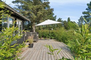 Eine Holzterrasse mit weißem Sonnenschirm, Kübelpflanzen und Grillplatz.