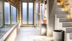 Un intérieur moderne avec un mur en pierre, une porte en verre donnant sur un balcon et un escalier orné d’un pot en cuivre posé sur un piédestal.