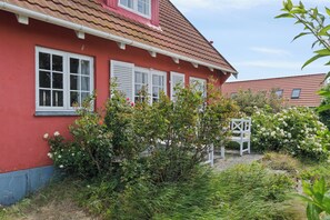 Un bâtiment rouge avec des fenêtres et des volets blancs, entouré de verdure et agrémenté d’un banc blanc.