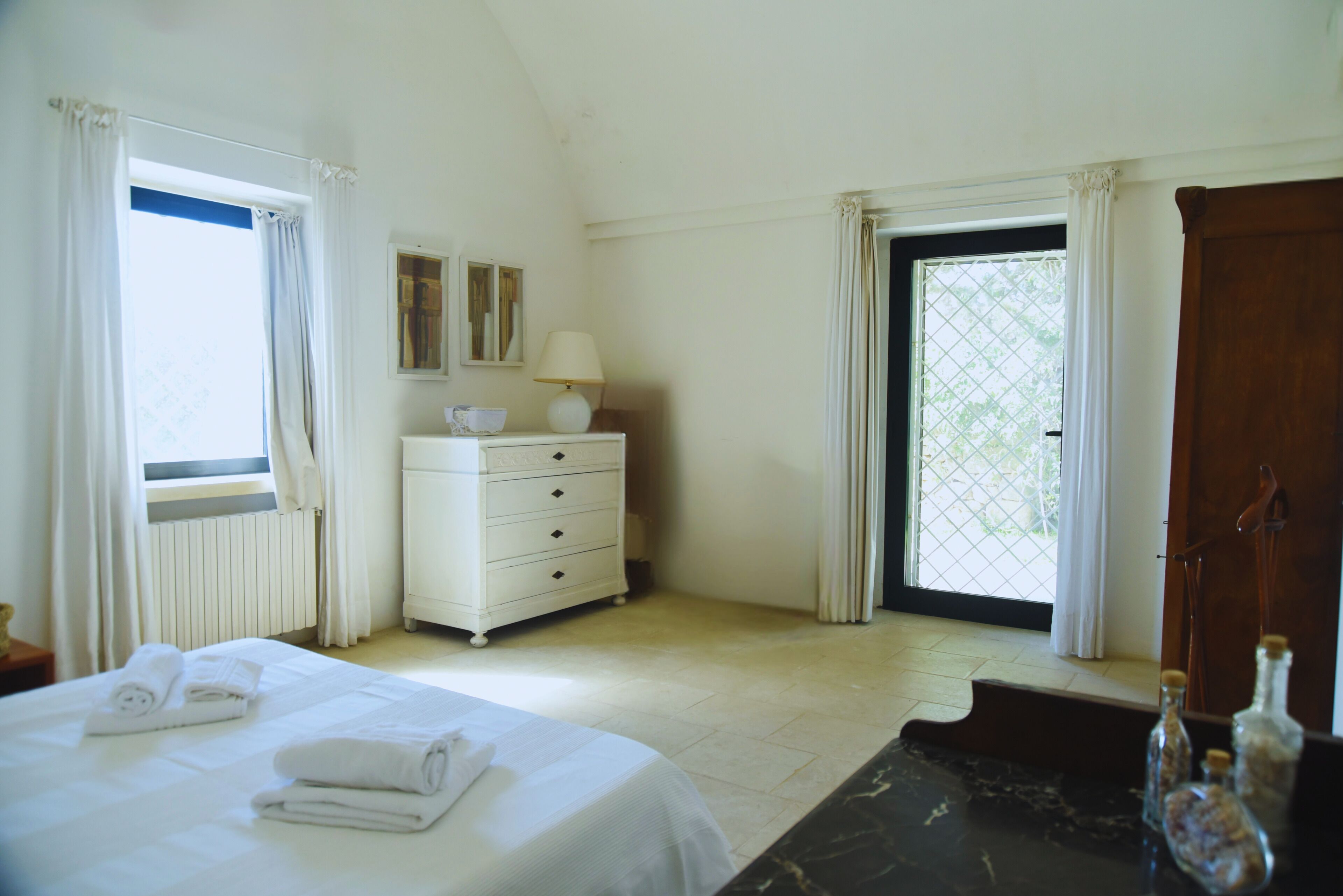 A hotel room with a bed, a dresser, a window with curtains, a lamp, and a view of greenery outside.
