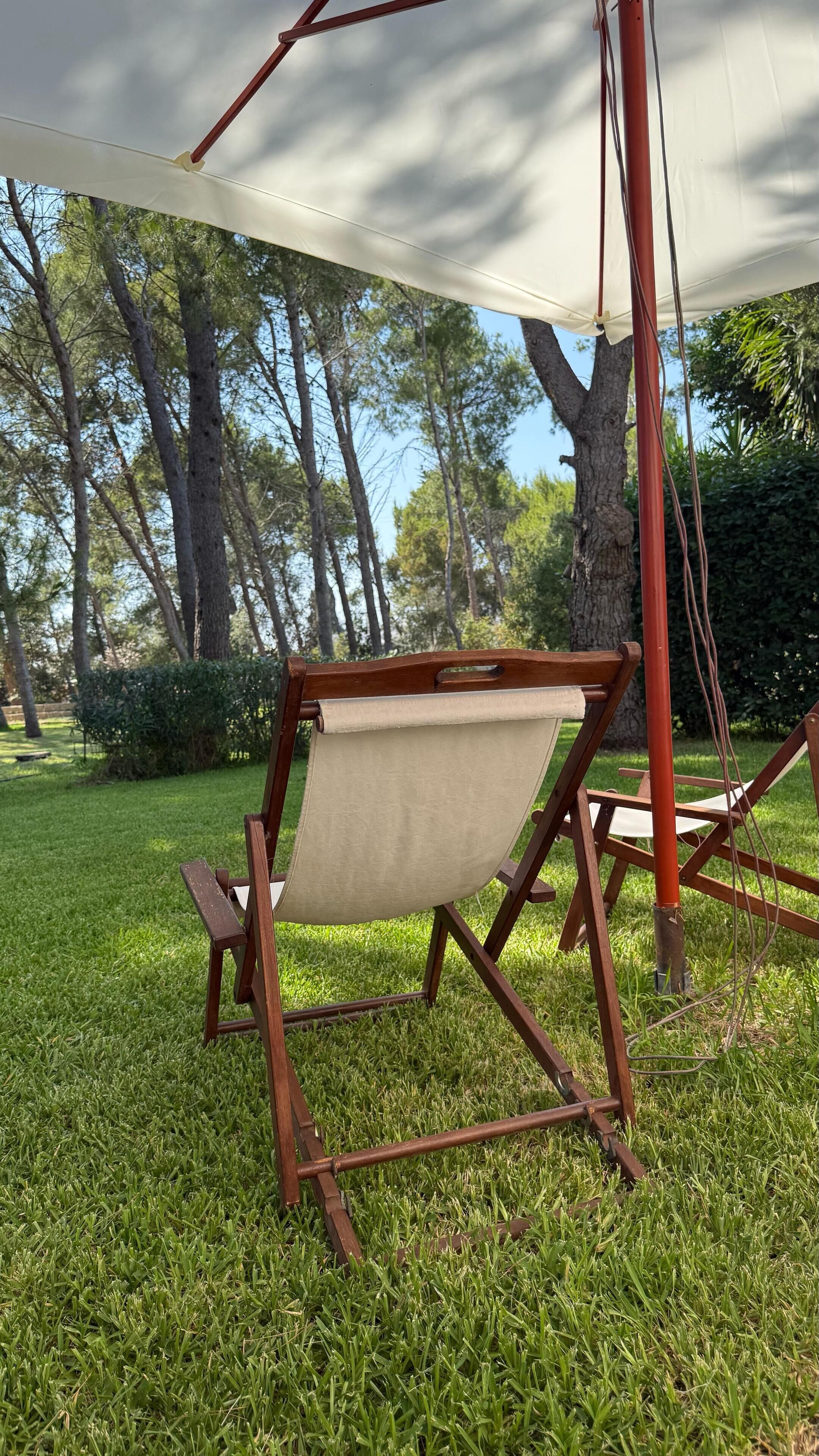 Une chaise en bois avec un dossier en toile, placée sous un auvent blanc dans un espace extérieur verdoyant, avec des arbres en arrière-plan.