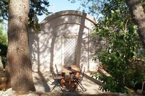 Un espacio al aire libre cubierto para comer con una mesa y sillas, rodeado de árboles y un muro de piedra.