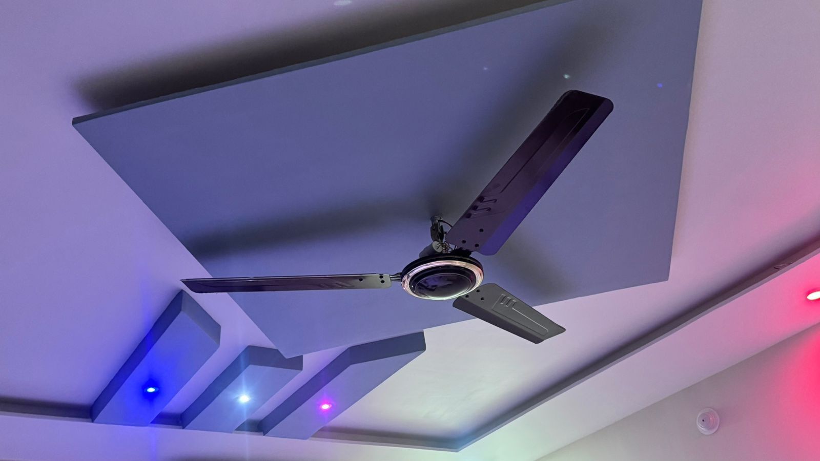 A ceiling fan with three blades, a central light fixture, and a smoke detector.