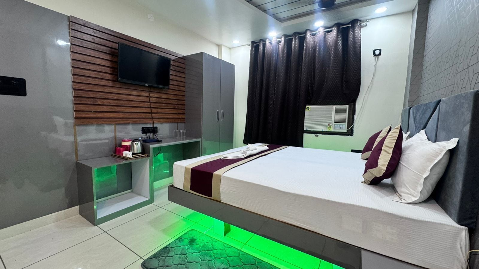 Una habitación de hotel moderna con cama, televisor montado en una pared de paneles de madera, un pequeño escritorio con hervidor y suelo iluminado en verde.