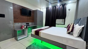 A modern hotel room with a bed, a TV mounted on a wooden panel wall, a small desk with a kettle, and a green-lit floor.