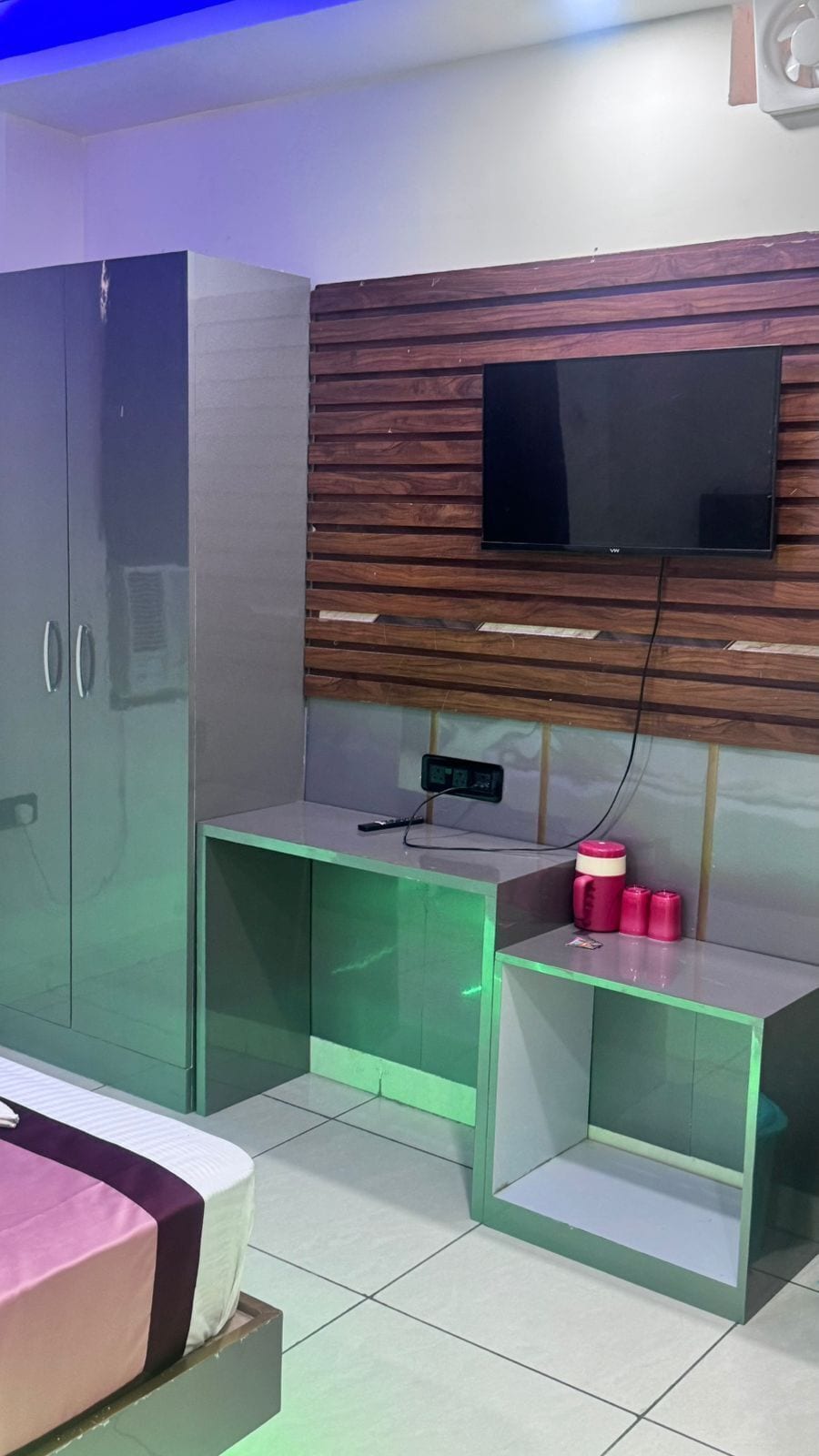 Una habitación con cama, armario, televisor montado en una pared de paneles de madera y un pequeño escritorio con toma de corriente y contenedores rosas.