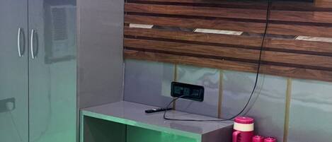 Una habitación con cama, armario, televisor montado en una pared de paneles de madera y un pequeño escritorio con toma de corriente y contenedores rosas.