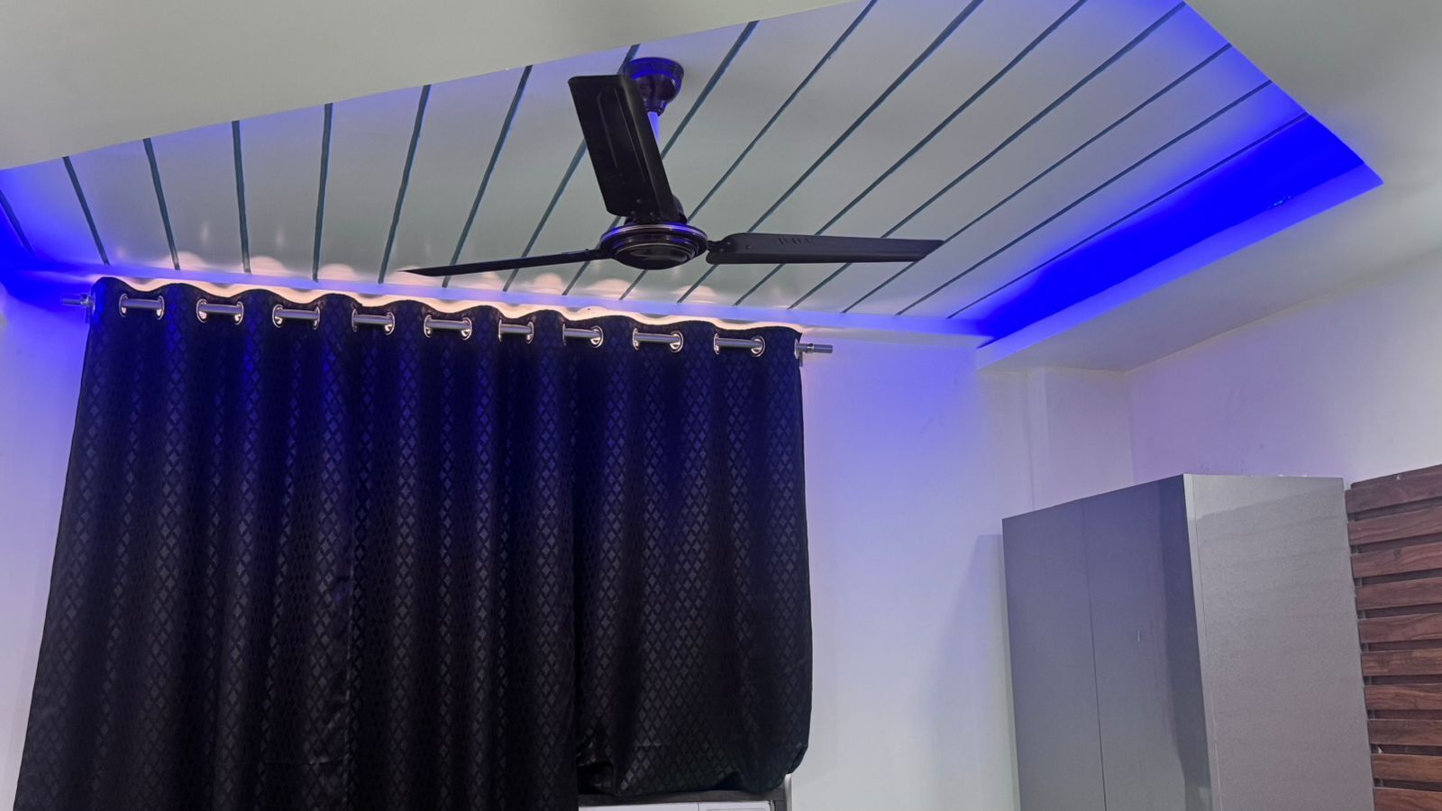 Un ventilador de techo con iluminación azul, una cortina negra y una pared blanca con panel de madera.