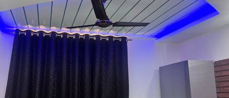 Un ventilador de techo con iluminación azul, una cortina negra y una pared blanca con panel de madera.