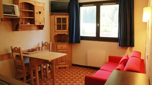 A compact room with a wooden dining table, chairs, a red sofa, a small cabinet with a microwave, and a window with curtains. - Apartment for 2 with mountain views (Châtel)