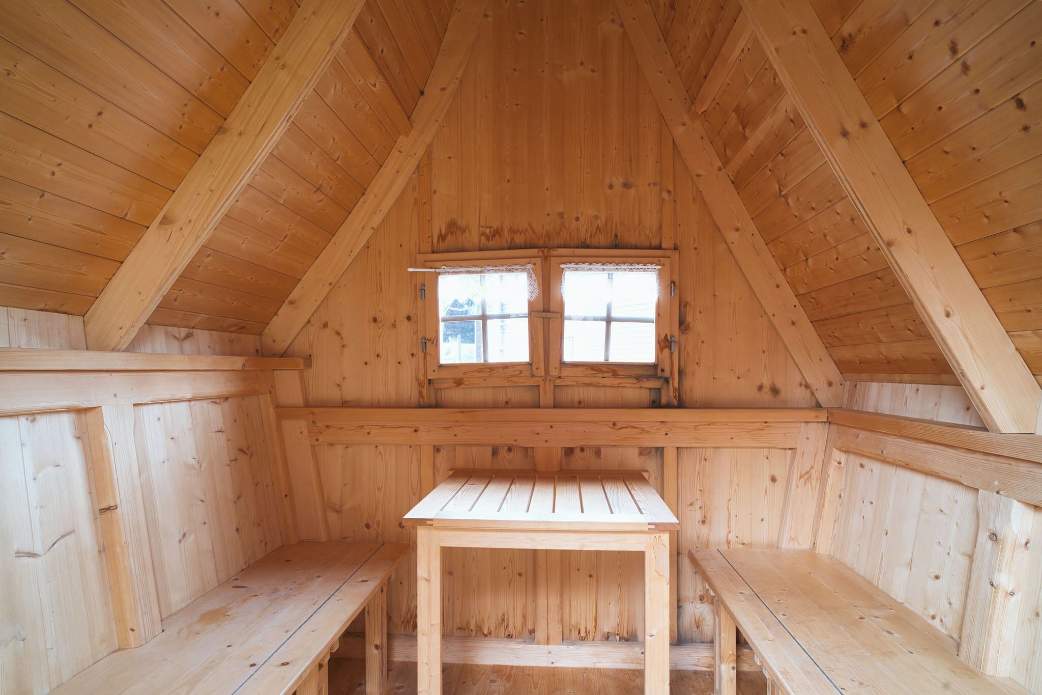 An attic room with wooden walls and a sloped ceiling. There is a small table and benches along the walls.
