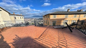 A rooftop terrace with terracotta tiles, a metal railing, and a view of a cityscape and mountains in the distance. - 6 Guests apt With Terrace in Pedestrian Historic Centre of Spoleto (Spoleto)