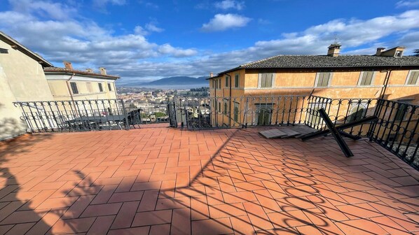 A rooftop terrace with terracotta tiles, a metal railing, and a view of a cityscape and mountains in the distance.