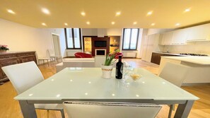 Une cuisine moderne avec des meubles blancs, un coin repas doté d’une table en verre et un salon avec un fauteuil rouge et une cheminée.