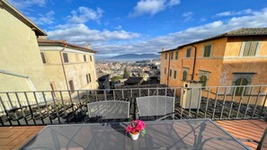 Eine Terrasse mit Metallgeländer, einem Blumentopf und Blick auf eine Stadtlandschaft mit Gebäuden und Bergen in der Ferne.