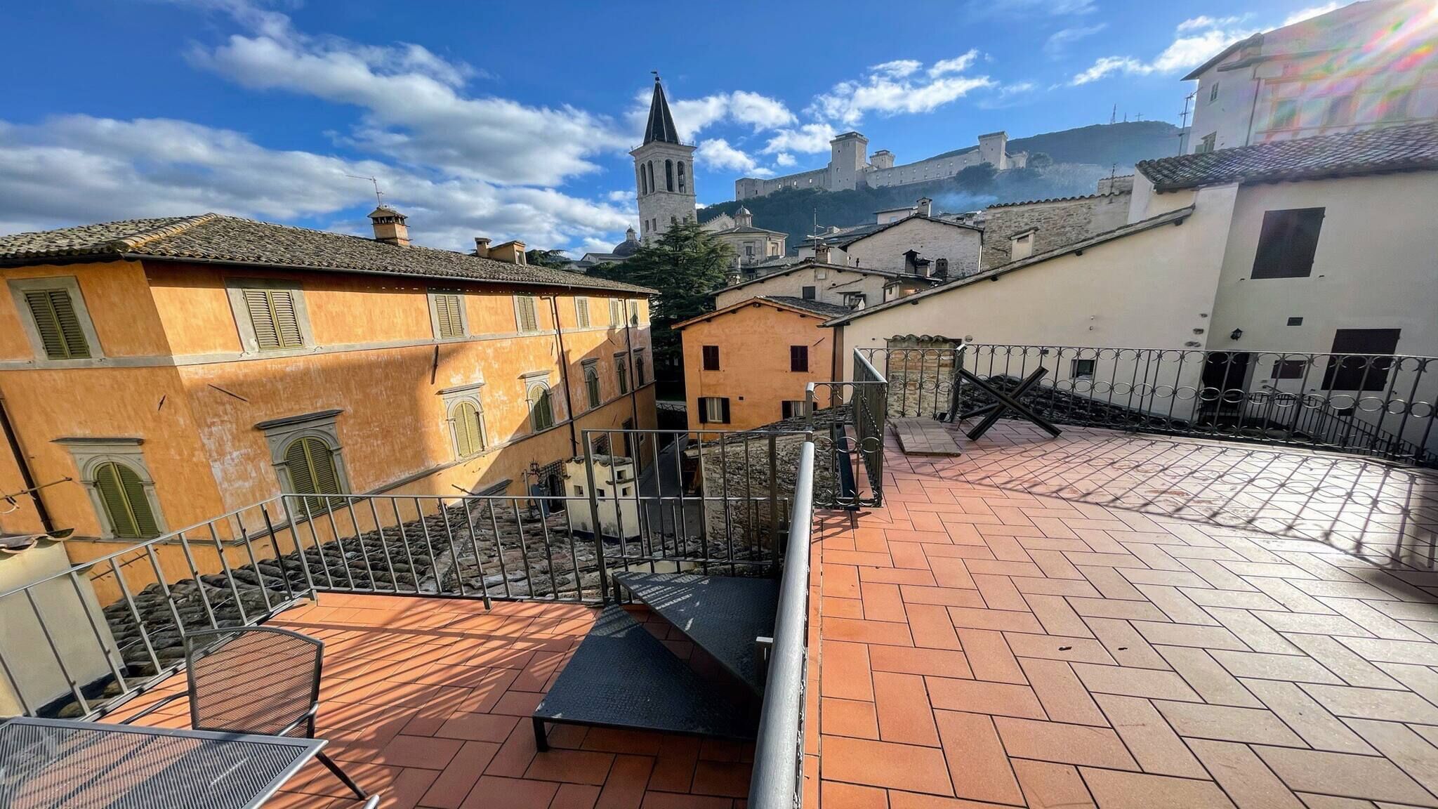 A rooftop terrace with a metal railing, a staircase, and a view of historic buildings and a church tower.