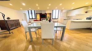 Una cocina moderna con armarios blancos, un comedor con mesa de vidrio y sillas blancas, y piso de madera.
