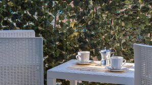 A white table with two white cups and a silver teapot on a woven placemat, set against a backdrop of green foliage. - VillaRealeFlats (Lecce)